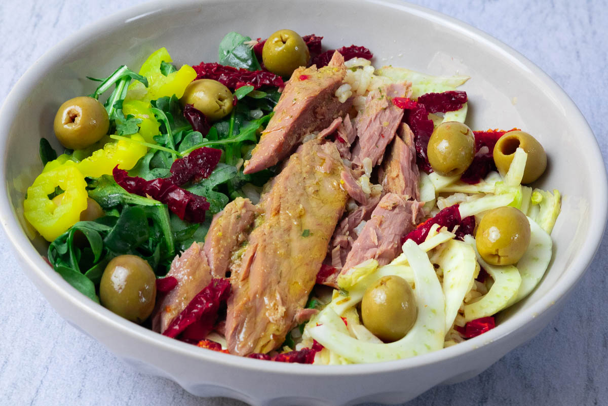 Tuna and rice salad bowl with olives, arugula, sun-dried tomatoes, and pepper rings in a bowl on a light background.