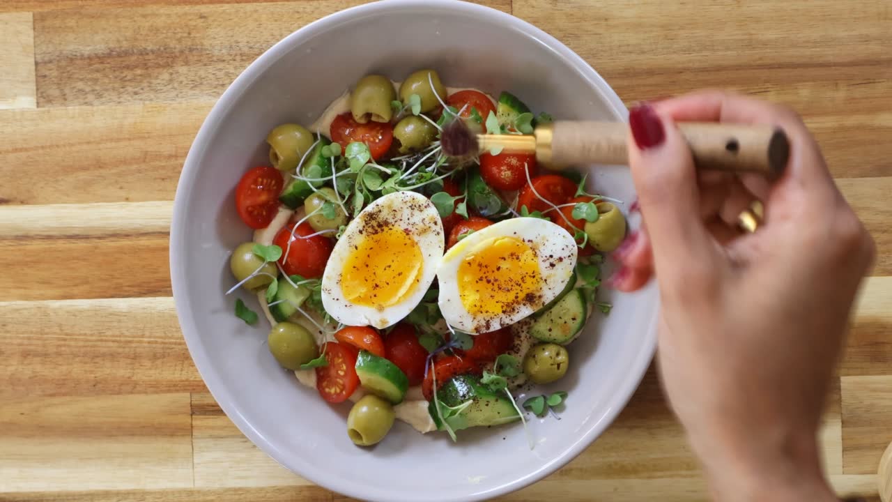 Bowl of salad with soft-boiled eggs, tomatoes, olives, and greens on a wooden table, seasoned by a hand.