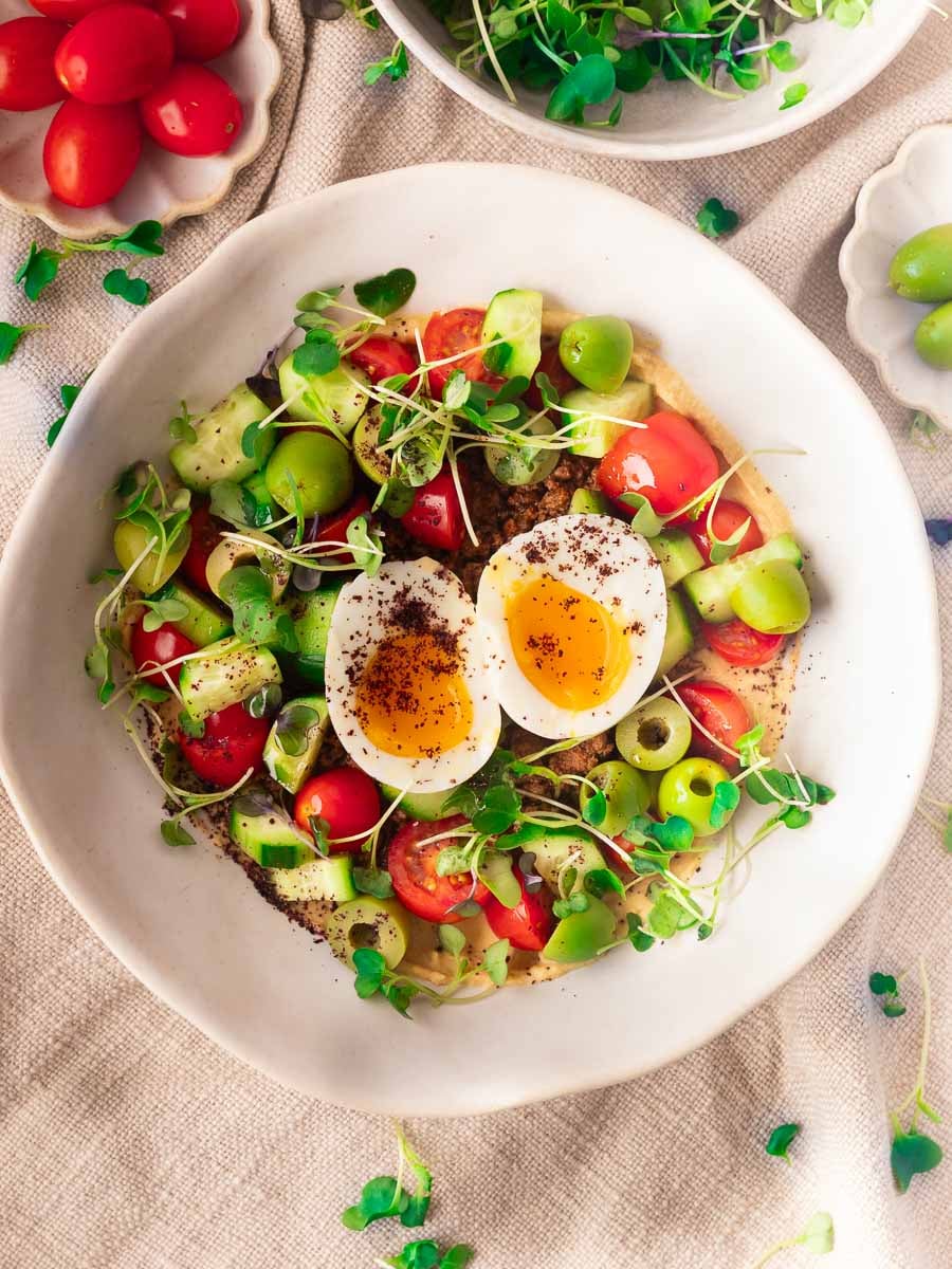 Mediterranean hummus bowl with halved boiled egg, cherry tomatoes, cucumbers, green olives, microgreens on a textured cloth.
