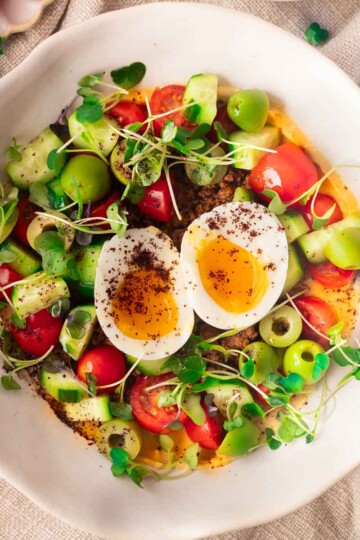Mediterranean hummus bowl with halved boiled egg, cherry tomatoes, cucumbers, green olives, microgreens on a textured cloth.