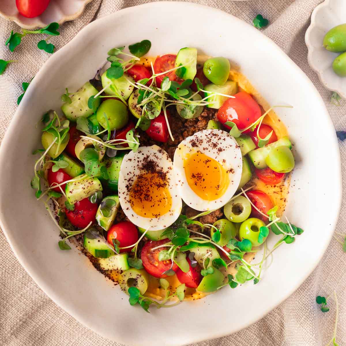 Mediterranean hummus bowl with halved boiled egg, cherry tomatoes, cucumbers, green olives, microgreens on a textured cloth.