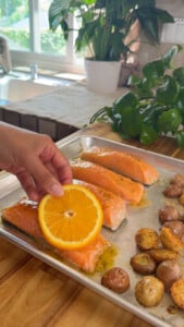 Hand placing orange slice on seasoned salmon fillets and potatoes on a baking tray.