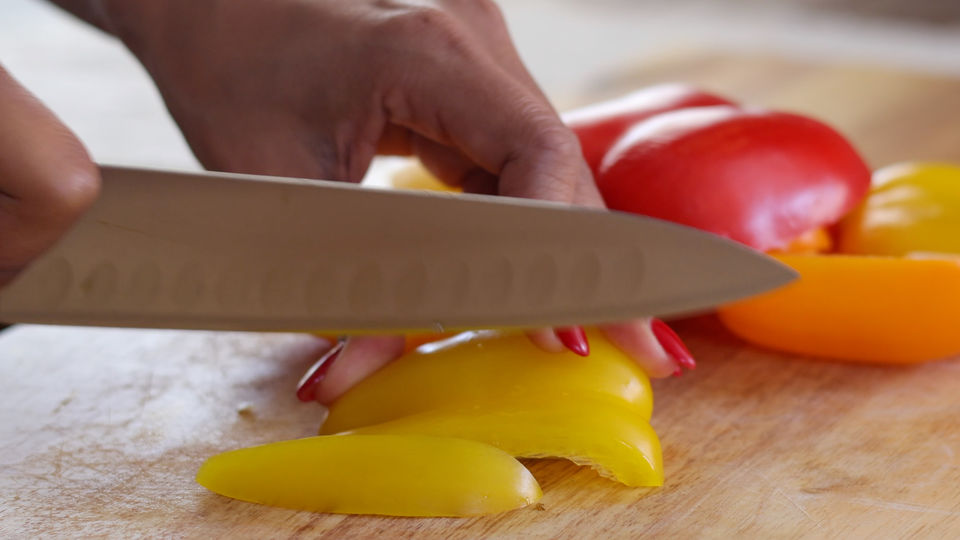 Close-up of a hand slicing yellow bell pepper on a wooden cutting board, with red and orange peppers nearby.
