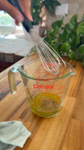 Whisk mixing oil and spices in a Pyrex measuring cup on a wooden counter.