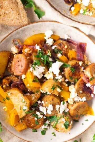 Plate of roasted potatoes, sausage, yellow peppers, red onions, and feta cheese, garnished with parsley, with bread.