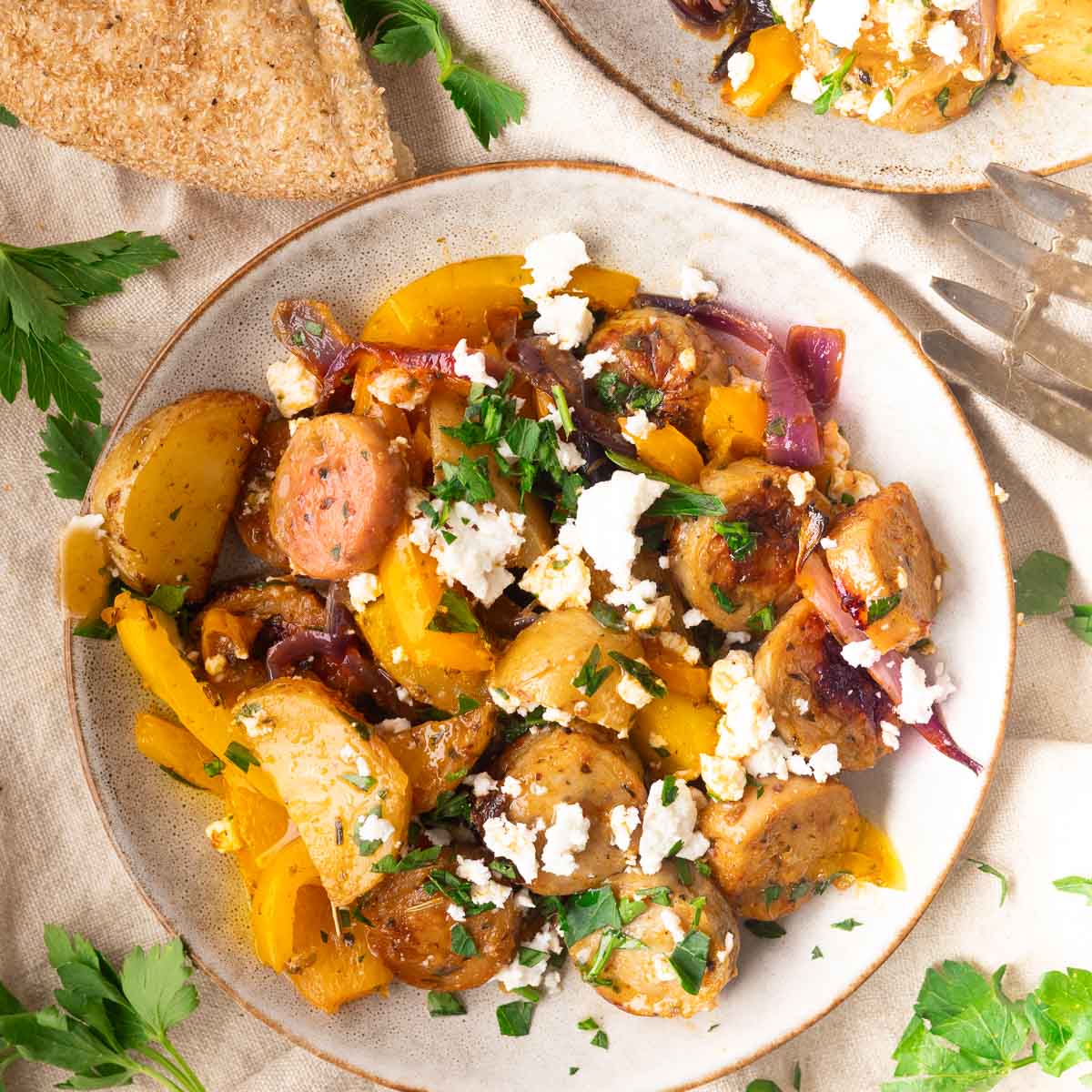 Plate of roasted potatoes, sausage, yellow peppers, red onions, and feta cheese, garnished with parsley, with bread.