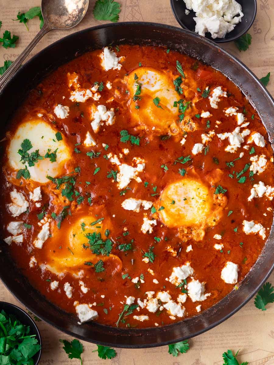 Shakshuka in a black skillet with poached eggs, tomato sauce, herbs, and cheese on a table with a spoon.