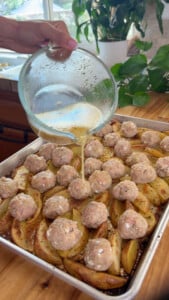 Pouring sauce over meatballs and potato wedges on a baking tray.