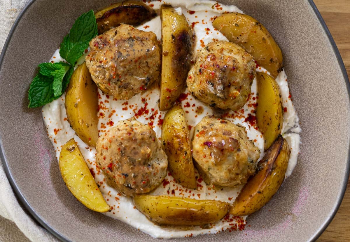Meatballs and roasted potatoes on yogurt sauce with herbs in a bowl.