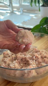 Hand shaping raw meatball over glass bowl with meat mixture on kitchen counter.