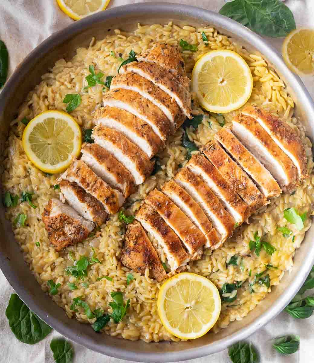 Sliced chicken breast on creamy orzo pasta with lemon slices and herbs in a skillet.