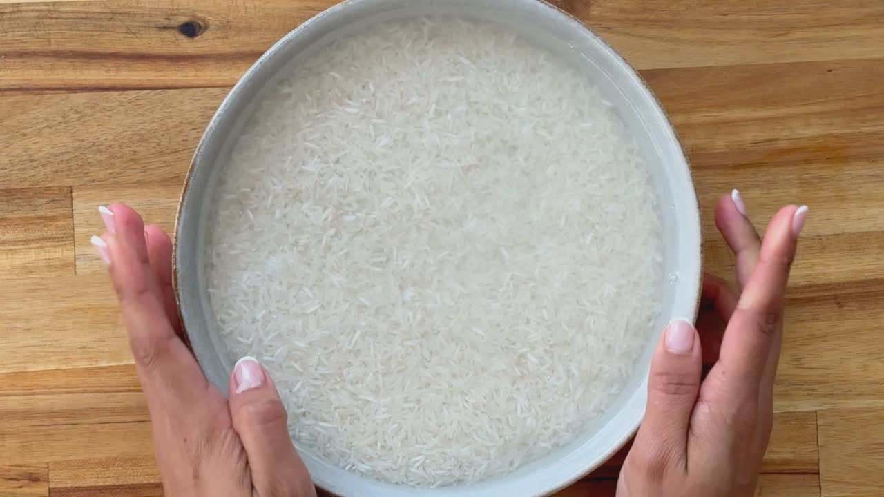 Hands holding a bowl of rice soaking in water on a wooden countertop.