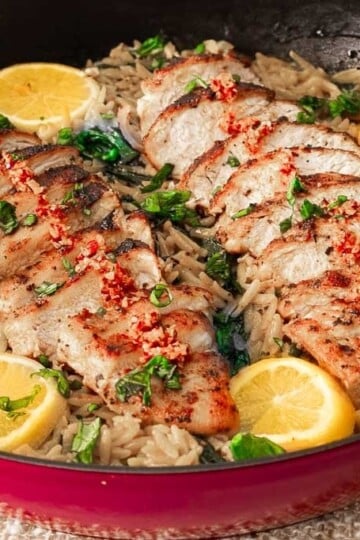 Grilled chicken with lemon slices and herbs on a bed of orzo pasta in a skillet.