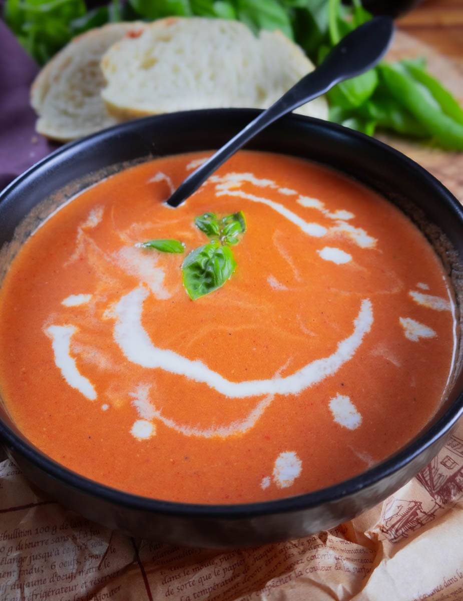 Roasted cherry & heirloom tomato soup garnished with basil in a black bowl, served with sliced bread in the background.