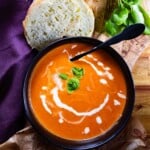 Roasted cherry & heirloom tomato soup garnished with basil leaves, served with rustic bread on a wooden table with a purple cloth.