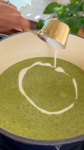 Pouring heavy cream in green soup in a pot.