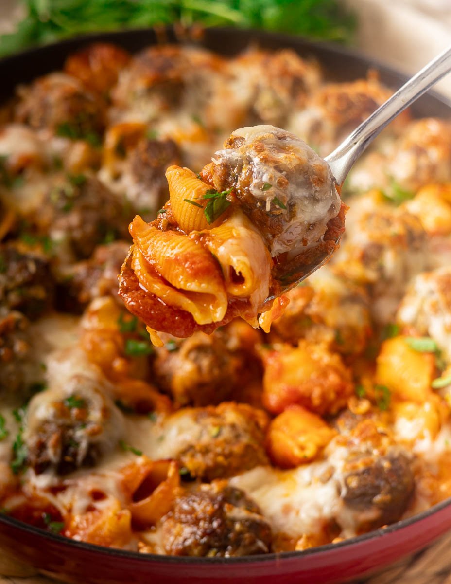 Cheesy baked meatballs and pasta with tomato sauce on a spoon over a skillet, garnished with herbs.