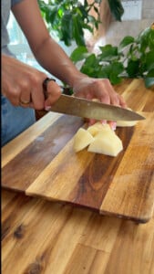 Hands chopping potato on a wooden cutting board with a chef’s knife in a kitchen.