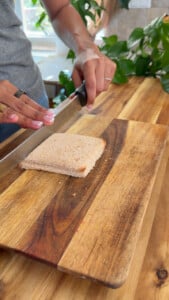 Person slicing bread on a wooden cutting board with a chef’s knife in a kitchen setting.