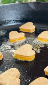 Mini heart cheese sandwiches cooking on a black griddle with butter melting in the pan.