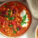 A bowl of hearty chili with beans, corn, tomatoes, sour cream, and parsley garnish on a marble countertop.