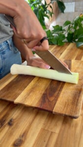 Person chopping a leek with a kitchen knife on a wooden cutting board in a kitchen.