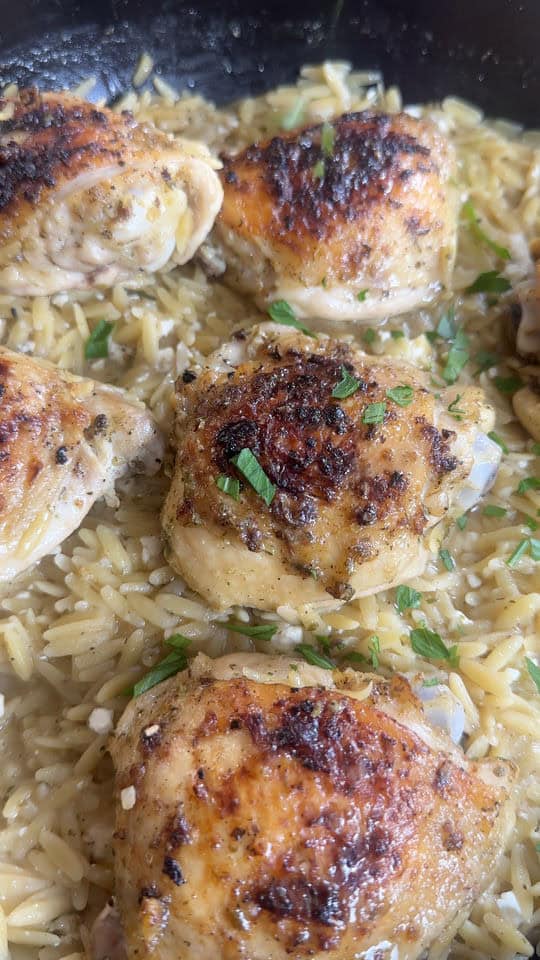 Close-up of browned chicken thighs with orzo pasta, garnished with fresh herbs in a skillet.