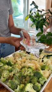 Hands preparing garlic over mixed vegetables in an aluminum tray.