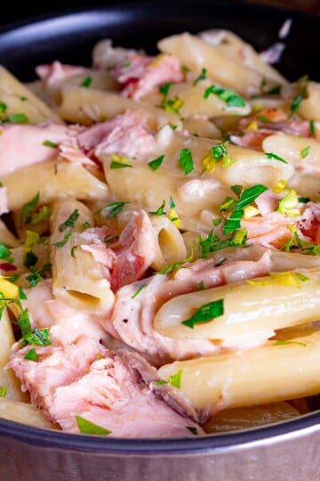 Creamy penne pasta with chunks of salmon and fresh parsley in a black skillet.