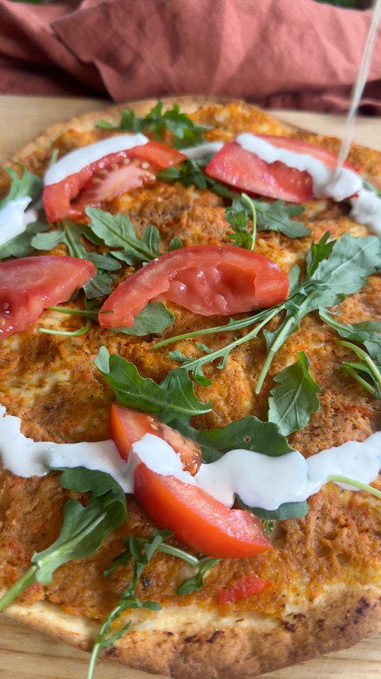 Flatbread topped with tomato slices, arugula, and drizzled yogurt sauce on a wooden board.