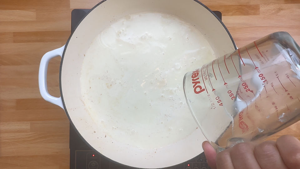 Pouring liquid from a measuring cup into a white pot on a stovetop.