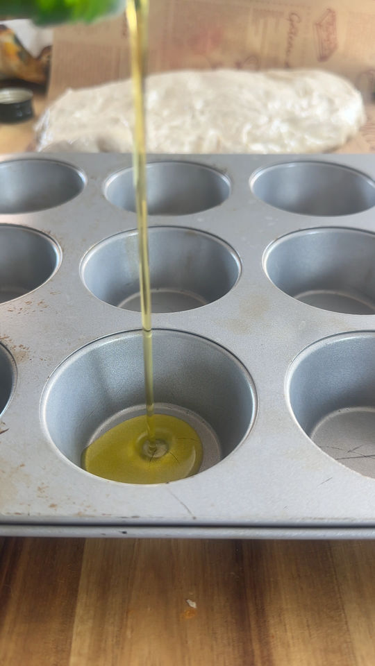 Pouring olive oil into a muffin tin cup, with dough wrapped in plastic on the table.