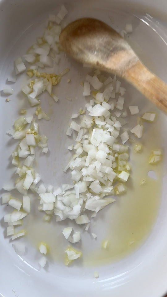 Chopped onions and garlic in oil with a wooden spoon in a white pan.