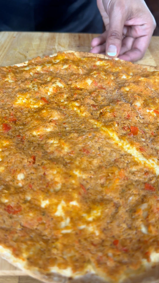 Close-up of a hand holding lahmacun with a minced meat topping on a wooden board.