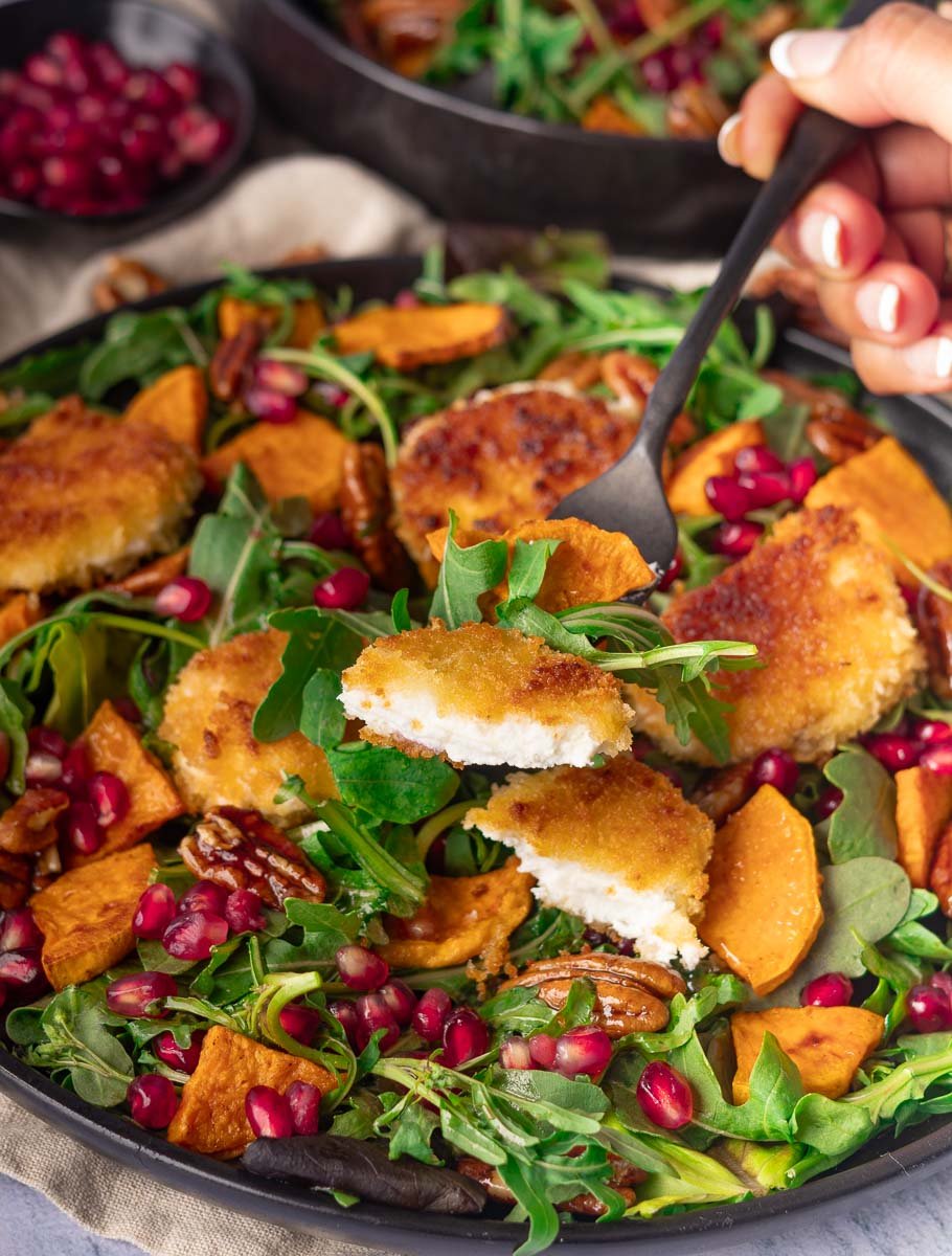 Crispy goat cheese salad with arugula salad with breaded goat cheese, sweet potatoes, pecans, and pomegranate seeds being forked.