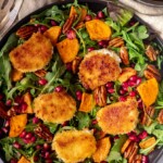 Crispy goat cheese salad with fried goat cheese, arugula, sweet potatoes, pecans, and pomegranate seeds in a black bowl.