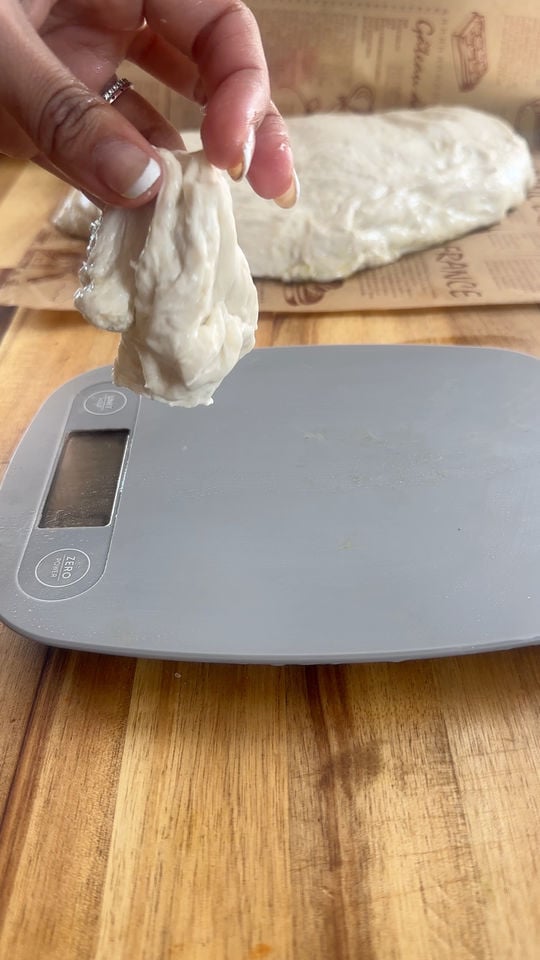Hand holding dough piece over digital kitchen scale on wooden surface.