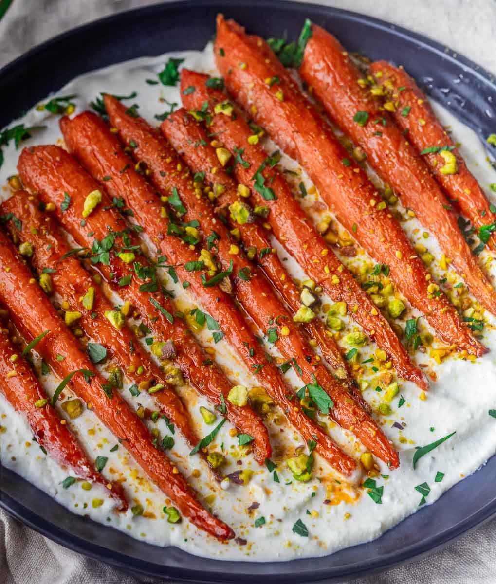 Roasted Harissa Maple  Carrots on whipped feta, garnished with pistachios and herbs, served on a dark oval plate.