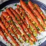 Roasted Harissa Maple Carrots, chopped pistachios and herbs on creamy sauce in a dark dish.