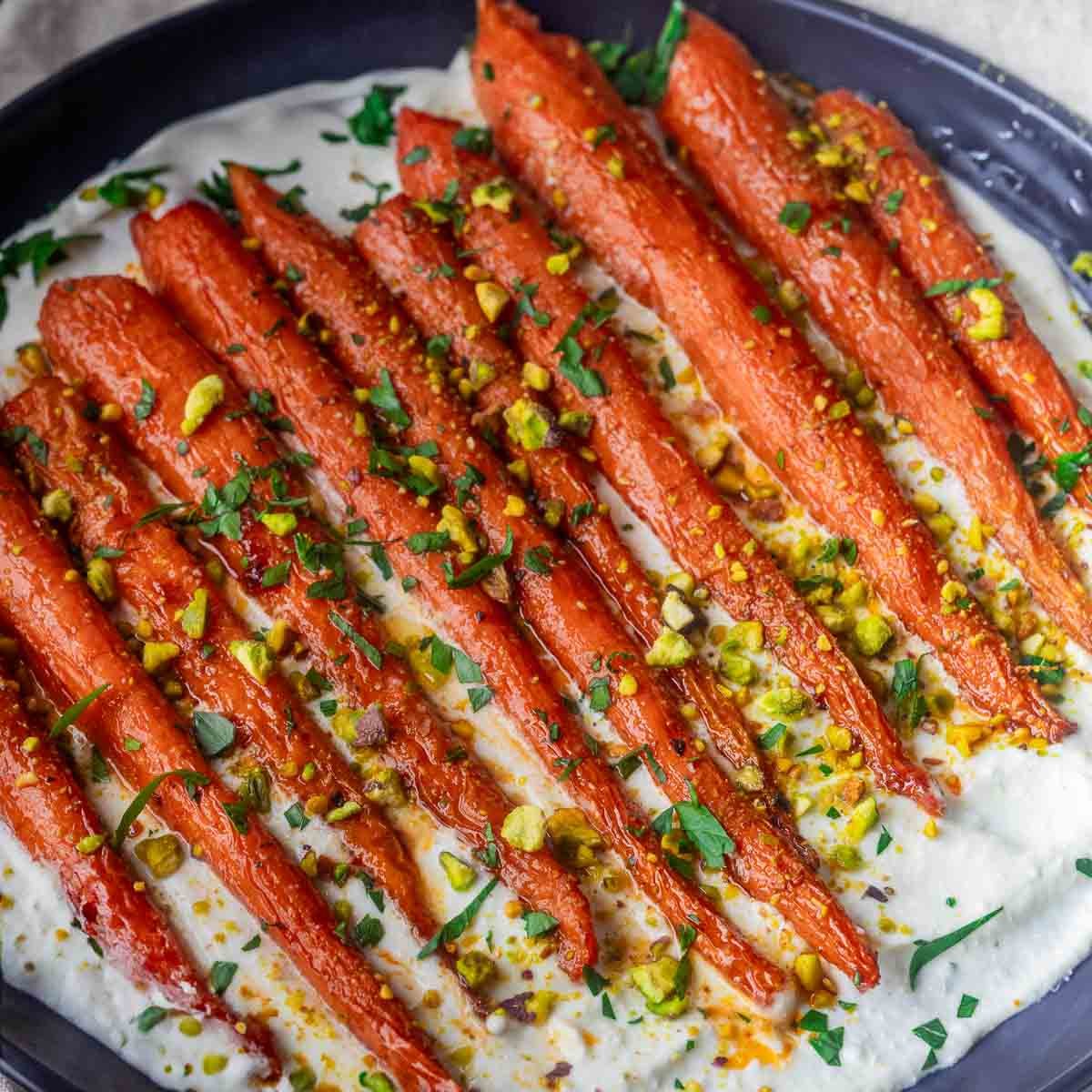 Roasted Harissa Maple Carrots, chopped pistachios and herbs on creamy sauce in a dark dish.