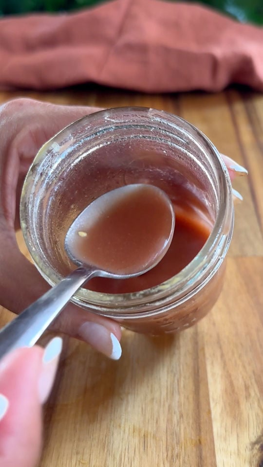Hand holding a spoon above a jar of salad dressing on a wooden table.