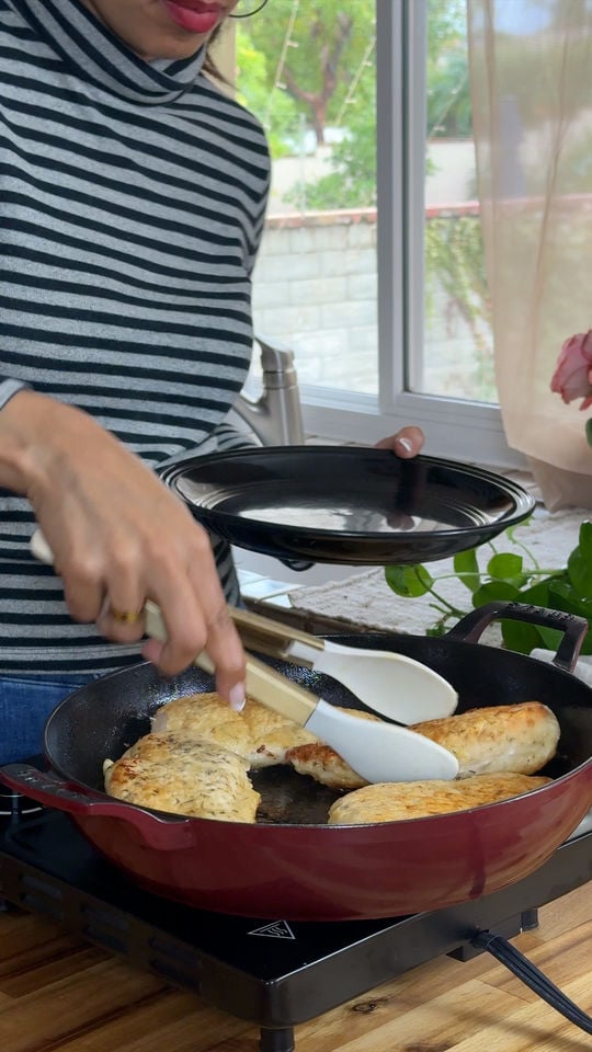Person in striped shirt cooking chicken breasts in a red skillet near a window.