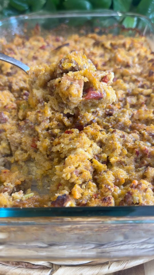Spoonful of homemade cornbread dressing with diced vegetables in a glass baking dish, garnished with herbs.