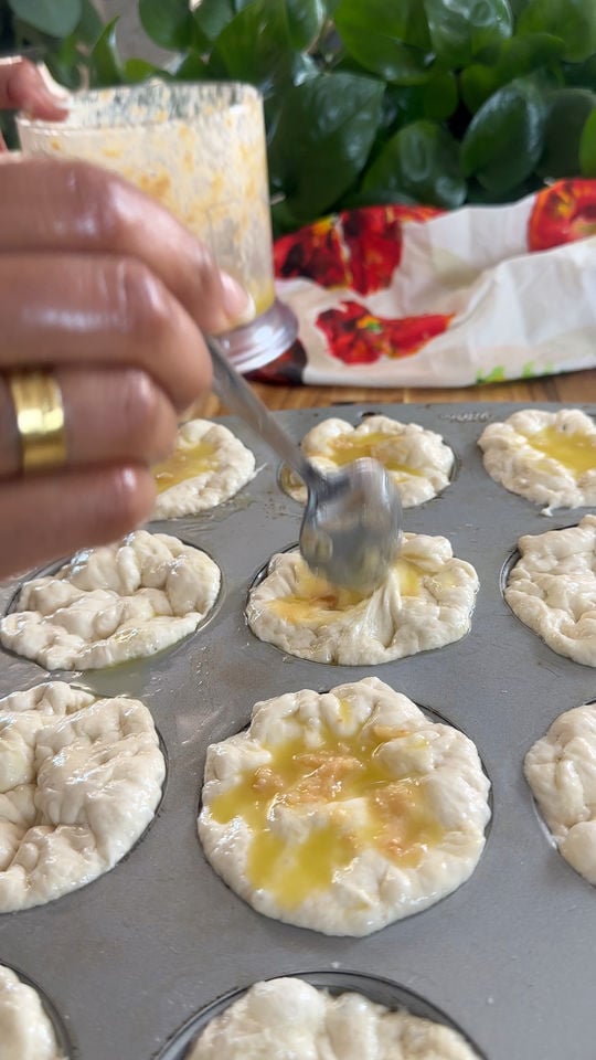 Hand spreading oil on raw pastry dough in muffin tin, with leafy plant and napkin in background.