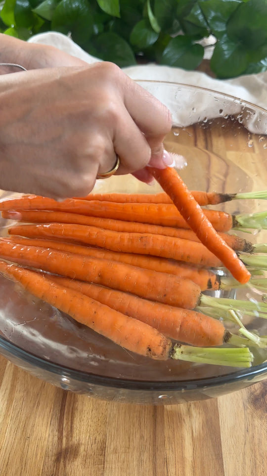 Hand washing fresh carrots in a glass bowl filled with water on a wooden surface.