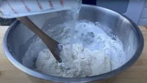 Pouring water into a mixing bowl of flour with a wooden spoon.