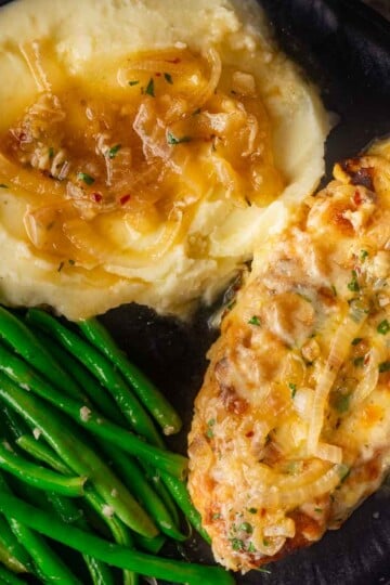 Cheesy chicken with caramelized onions, mashed potatoes, and green beans on a black plate.