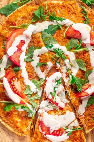 Lebanese chicken pizza with tomato slices, arugula, and white sauce drizzle on a wooden board.