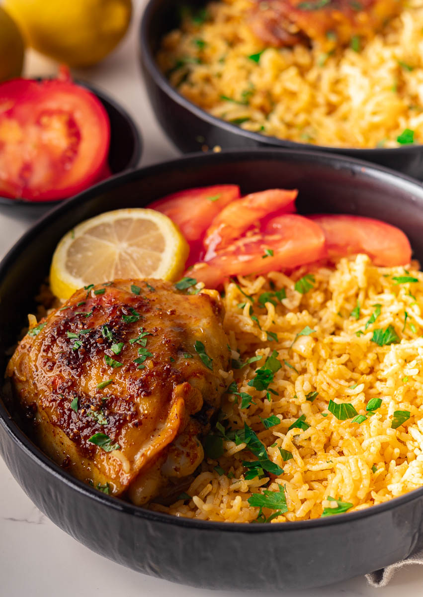 Mediterranean baked chicken thighs and rice with sliced tomatoes, and lemon garnish in a black bowl.