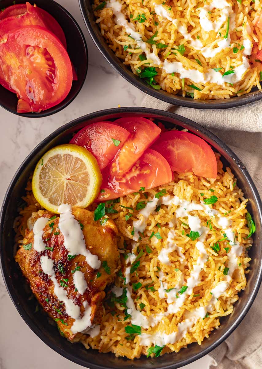 Mediterranean Baked Chicken Thighs and Rice with tomatoes, lemon, and sauce in black bowls on marble surface.